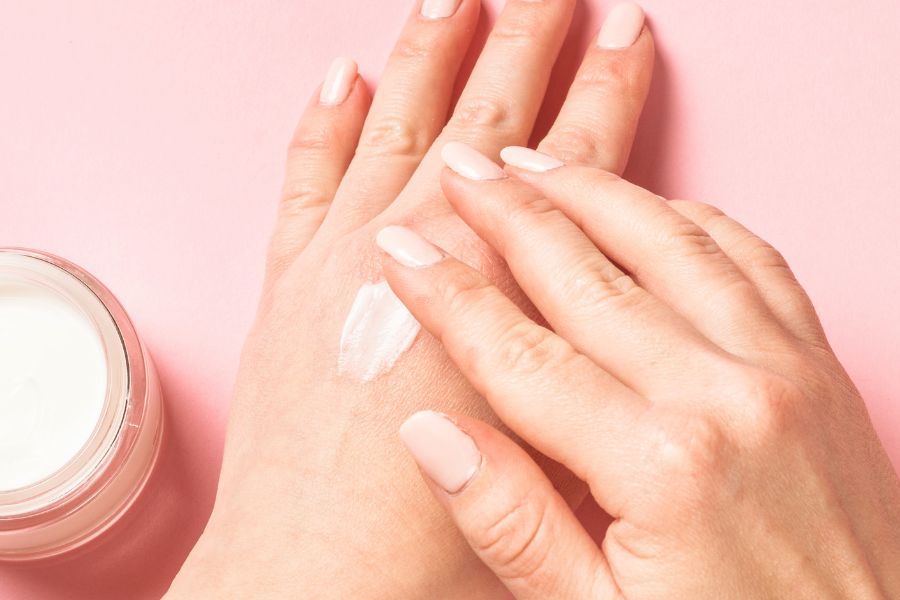 Une personne applique une crème hydratante blanche sur le dos de sa main sur un fond rose